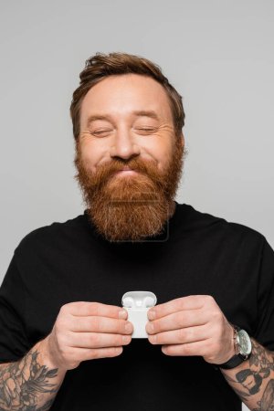 Photo for Pleased bearded man with closed eyes holding case with wireless headphones isolated on grey - Royalty Free Image