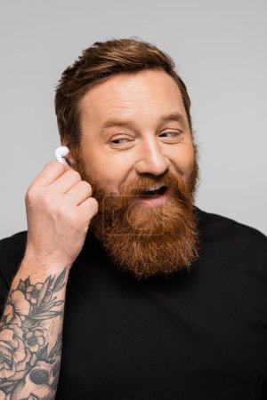 amazed and happy bearded man holding wireless earphone isolated on grey