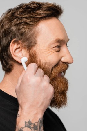 Foto de Vista lateral del hombre barbudo alegre sosteniendo auricular inalámbrico cerca de la oreja aislado en gris - Imagen libre de derechos