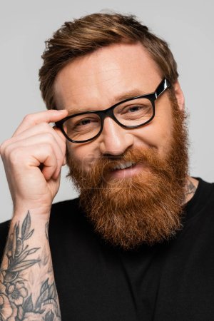 portrait of positive bearded man adjusting eyeglasses and smiling at camera isolated on grey