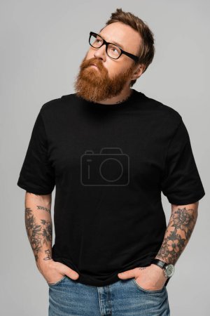 stylish bearded man in eyeglasses standing with hands in pockets of jeans and looking away isolated on grey