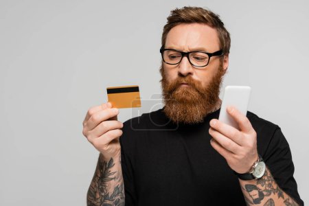 Foto de Thoughtful and stylish man in eyeglasses holding credit card and mobile phone isolated on grey - Imagen libre de derechos