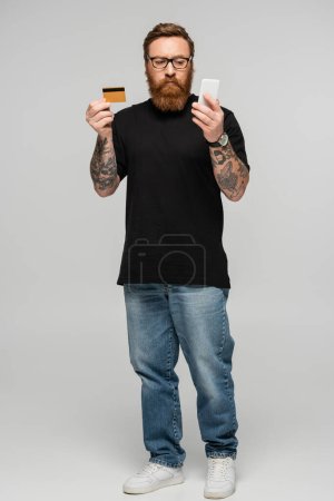 full length of thoughtful man in eyeglasses holding cellphone and credit card on grey background