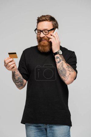 happy tattooed man in eyeglasses talking on mobile phone while holding credit card isolated on grey