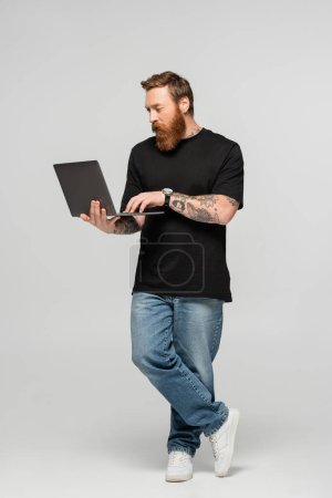 Foto de Longitud completa del hombre enfocado en jeans y camiseta negra usando portátil sobre fondo gris - Imagen libre de derechos