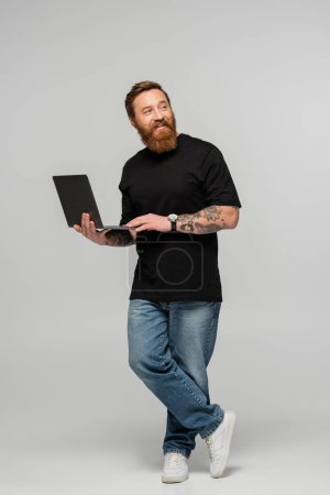 full length of positive bearded man in jeans standing with laptop and looking away on grey background