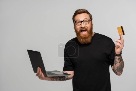 Foto de Amazed bearded man in eyeglasses holding laptop and showing credit card isolated on grey - Imagen libre de derechos