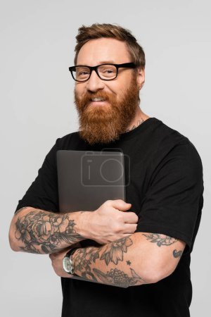 pleased bearded freelancer in eyeglasses holding laptop and smiling at camera isolated on grey