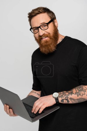 bearded freelancer in eyeglasses holding laptop and smiling at camera isolated on grey