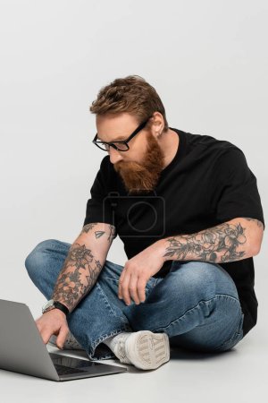 focused bearded freelancer in eyeglasses working on laptop while sitting with crossed legs on grey background