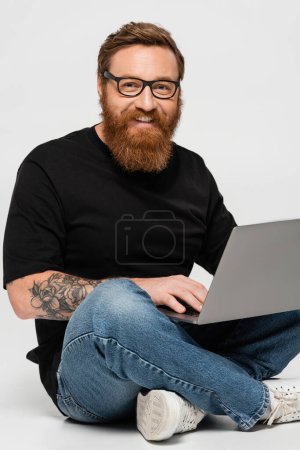 happy bearded freelancer holding laptop while sitting with crossed legs on grey background