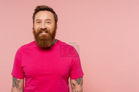 positive bearded and tattooed man in magenta t-shirt smiling at camera isolated on pink