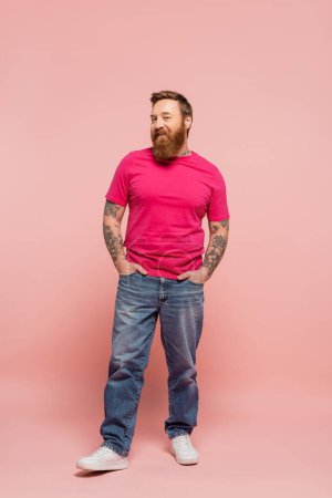 full length of happy bearded man in magenta t-shirt and jeans posing with hands in pockets on pink background