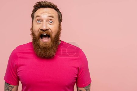 impressed bearded man with open mouth looking at camera isolated on pink