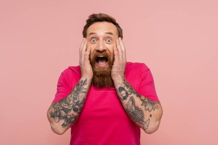 amazed bearded man in bright t-shirt touching face while looking at camera isolated on pink