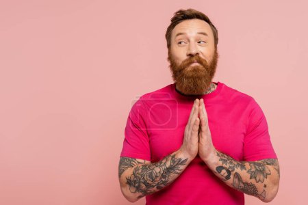 Photo for Smiling and tricky man in magenta t-shirt showing please gesture and looking away isolated on pink - Royalty Free Image
