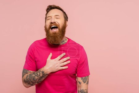 joyful bearded man in bright t-shirt touching chest and laughing with closed eyes isolated on pink