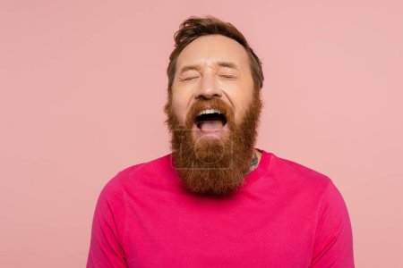 excited bearded man in magenta t-shirt laughing with closed eyes isolated on pink