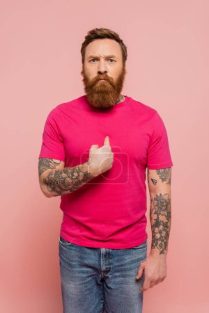 displeased bearded man in trendy t-shirt pointing with finger at himself isolated on pink