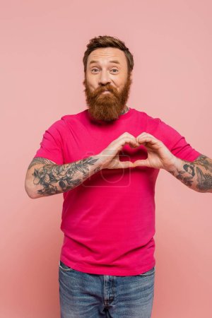 cheerful tattooed and bearded man in magenta t-shirt showing heart sign while looking at camera isolated on pink