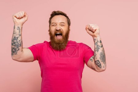 joyful bearded man with closed eyes showing triumph gesture and screaming isolated on pink