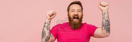 Téléchargez les photos : Excité tatoué homme avec les yeux fermés criant et montrant geste gagnant isolé sur rose, bannière - en image libre de droit
