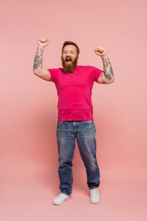 full length of trendy man in magenta t-shirt and jeans rejoicing and screaming on pink background