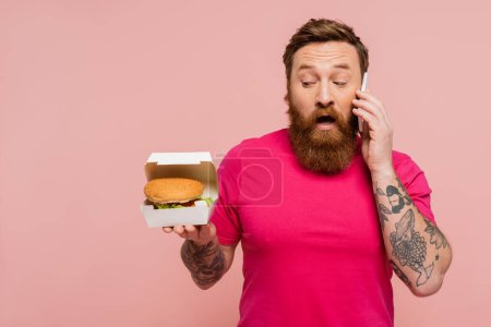hombre barbudo asombrado hablando por teléfono móvil y sosteniendo paquete de cartón con sabrosa hamburguesa aislada en rosa