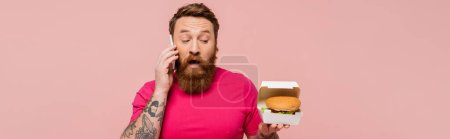 Aufgeregter Mann hält Kartonpackung mit leckerem Burger in der Hand, während er auf dem Smartphone mit rosa Banner spricht