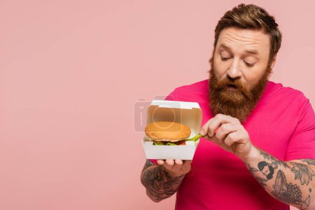 barbudo hombre tomando papas fritas de paquete de cartón con sabrosa hamburguesa aislado en rosa