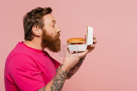 Téléchargez les photos : Vue latérale de l'homme tatoué barbu sentant hamburger savoureux dans le paquet de carton isolé sur rose - en image libre de droit
