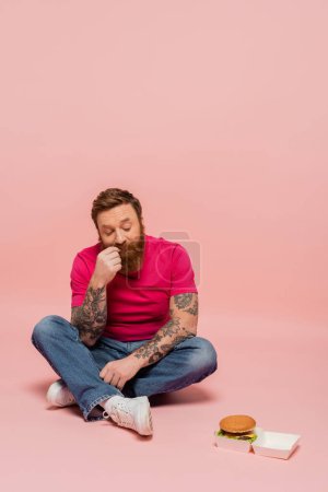 volle Länge des nachdenklichen Mannes in Jeans, der Kartonpackung mit Burger betrachtet, während er mit überkreuzten Beinen auf rosa Hintergrund sitzt