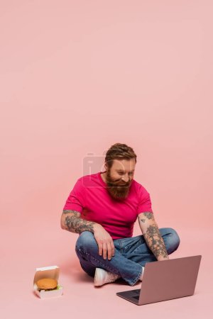 lächelnder bärtiger Freiberufler, der am Laptop arbeitet, während er mit gekreuzten Beinen in der Nähe der Packung mit Hamburger auf rosa Hintergrund sitzt