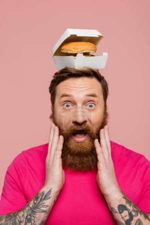 astonished man with hamburger in carton pack on head touching beard and looking at camera isolated on pink