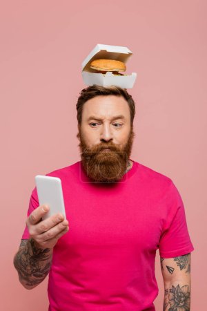 hombre barbudo reflexivo mirando el teléfono inteligente, mientras que de pie con sabrosa hamburguesa en la cabeza aislado en rosa