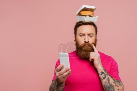 hombre barbudo reflexivo con hamburguesa en la cabeza tocando la cara y mirando el teléfono móvil aislado en rosa