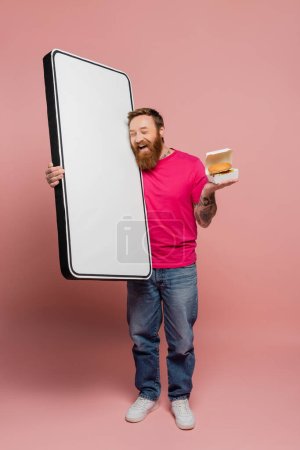 bearded man holding hamburger and huge template of mobile phone while laughing with closed eyes on pink background