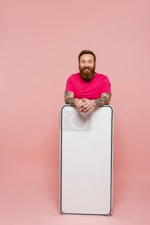 bearded and tattooed man in magenta t-shirt looking at camera near huge phone template on pink background