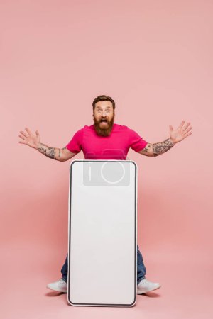 hombre tatuado emocionado mostrando gesto wow cerca de plantilla de teléfono móvil enorme sobre fondo rosa