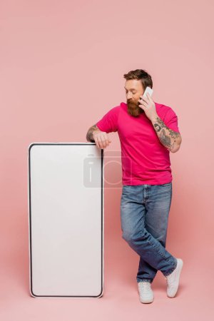 longitud completa del hombre barbudo en jeans hablando en el teléfono inteligente cerca del teléfono blanco maqueta sobre fondo rosa