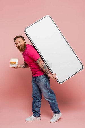 happy bearded man in magenta t-shirt and jeans carrying hamburger and white template of smartphone on pink background
