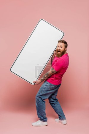 longitud completa de hombre barbudo excitado sosteniendo plantilla de teléfono enorme y mirando a la cámara en el fondo rosa