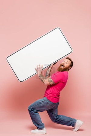 longitud completa del hombre asombrado mirando a la cámara mientras sostiene la plantilla de teléfono grande sobre fondo rosa