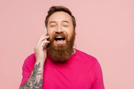 joyful bearded man with closed eyes talking on mobile phone and laughing isolated on pink