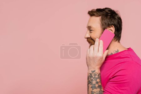 Téléchargez les photos : Vue latérale de l'homme joyeux en t-shirt magenta appelant sur téléphone mobile isolé sur rose - en image libre de droit