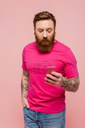 tattooed man in magenta t-shirt standing with hand in pocket of jeans and using smartphone isolated on pink