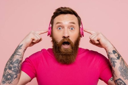 amazed bearded man in magenta t-shirt pointing at wireless headphones and looking at camera isolated on pink