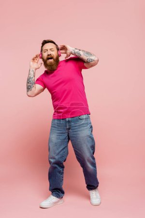 Téléchargez les photos : Full length of astonished man with closed eyes listening music in wireless headphones and singing on pink background - en image libre de droit