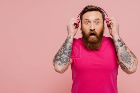 Foto de Surprised tattooed man with open mouth holding wireless headphones while looking at camera isolated on pink - Imagen libre de derechos