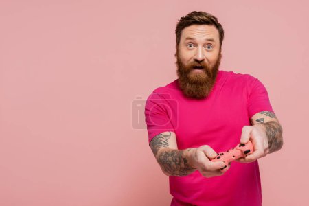 Foto de KYIV, UCRANIA - 12 DE DICIEMBRE DE 2022: hombre barbudo excitado en camiseta magenta jugando videojuegos aislados en rosa - Imagen libre de derechos
